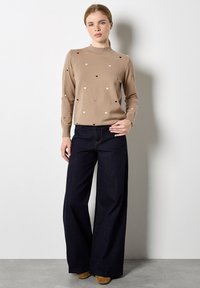 Tan trui met kleine, multikleurige hartpatronen; geribbelde halslijn en boorden. Gemaakt in combinatie met losse, donkere spijkerbroek en beige schoenen.