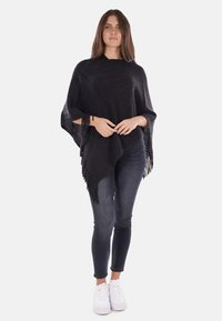 Poncho nero a maglia con una texture a coste, dotato di orli frangiati. Abbinato a jeans scuri e sneaker bianche.