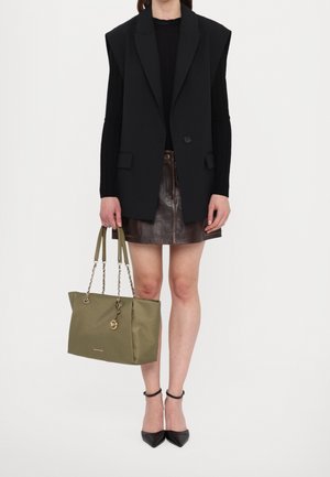 Mujer con blazer negro de gran tamaño, top negro de manga larga, minifalda de cuero marrón, tacones negros, sosteniendo un bolso de mano verde oliva con asas de cadena.