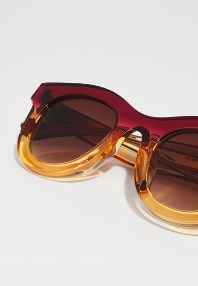 Lunettes de soleil avec un dégradé rouge et orange, à monture surdimensionnée et arrondie. Bord inférieur transparent et verres teintés brun foncé. Notez le logo sur la branche.