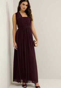 Langes, ärmelloses, lila Kleid mit tailliertem Oberteil, geraffter Textur und fließendem Rock. Die Modells hält eine runde, verzierte Clutch.