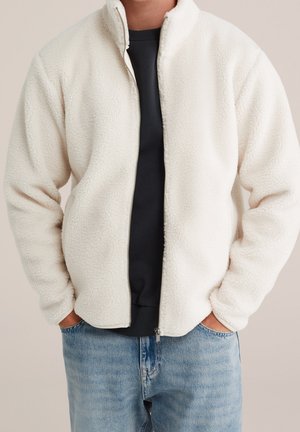 Mand iført en cremefarvet fleecetrøje over en sort skjorte og blå jeans, med hænderne stikket ned i jakkelommerne.