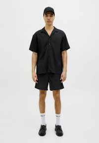 Chemise noire à manches courtes et shorts assortis en tissu texturé avec des rayures verticales. Porteé avec une casquette, des chaussettes blanches et des chaussures noires.