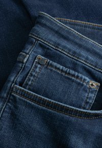 Donkerblauwe spijkerbroek met een voorvakdetail, zichtbare stiksels en metalen klinknagels. De stof heeft een licht gestructureerde afwerking.