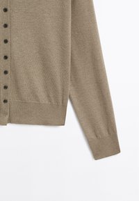 Manche et côté d’un cardigan en maille couleur taupe, avec des boutons noirs et des bords-côtes aux poignets et à l’ourlet sur fond blanc.