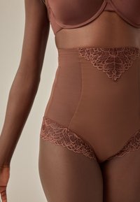 Femme portant un slip gainant marron taille haute avec des détails en dentelle et un soutien-gorge assorti, montrée du milieu du torse jusqu'aux cuisses.