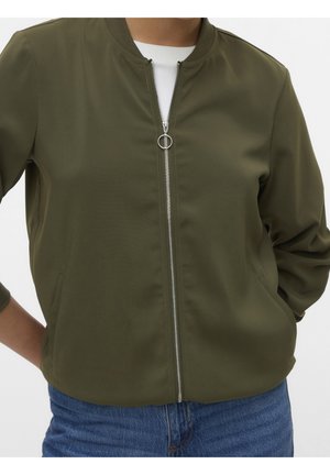 Personne portant une veste bomber zippée vert olive avec col et poignets côtelés sur une chemise blanche, associée à un jean bleu.