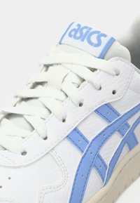 Λευκά αθλητικά παπούτσια με ανοιχτό μπλε ρίγες και κορδόνια, εμφανίζοντας το λογότυπο "asics" στην γλώσσα σε μπλε γράμματα, κοντινή θέα.