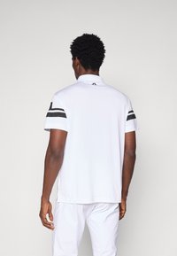 Camisa polo branca de manga curta com colarinho, apresentando detalhes em preto nas mangas e um logótipo nas costas perto do colarinho.