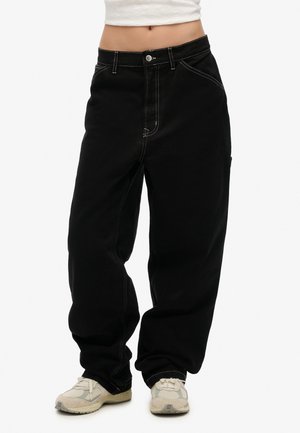 Black baggy jeans with white stitching, high-rise waist, button closure, and side pockets, paired with light-colored athletic shoes.