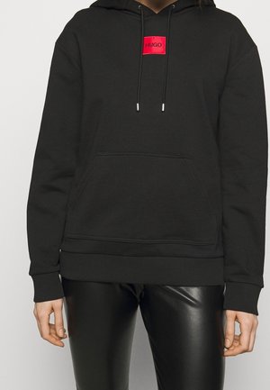 Personne portant un sweat à capuche noir avec un patch logo rouge "HUGO" et un pantalon en cuir noir, debout devant un fond uni clair.