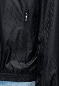 Μαύρο μπουφάν αδιάβροχο με διακριτικό γεωμετρικό σχέδιο, τσέπη με φερμουάρ, ελαστική μέση και απαλό, ελαφρύ ύφασμα.