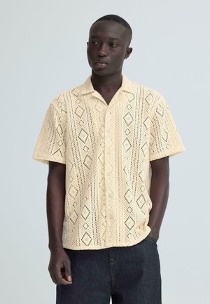 Hombre con camisa de crochet de manga corta color crema con patrones de diamantes y jeans azul oscuro, de pie contra un fondo liso gris claro.