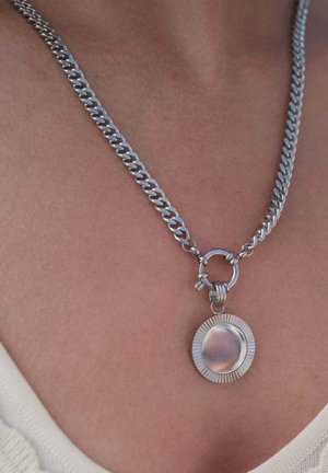 Silberne Halskette mit rundem, geriffeltem Anhänger, getragen auf nackter Haut über einem cremefarbenen Oberteil.