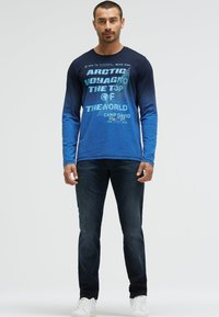 Langärmeliges blaues T-Shirt mit Farbverlauf verfügt über weißen Grafiktext, kombiniert mit dunklen Jeans und weißen Sneakern, getragen von einem stehenden männlichen Modell.