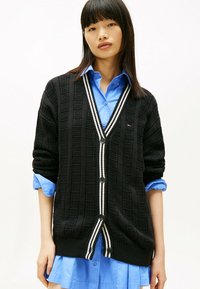 Zwarte gebreide cardigan met een gemêleerd patroon, voorzien van een V-hals, witte gestreepte accenten en zwarte knopen. Draag over een blauwe overhemd met knopen.