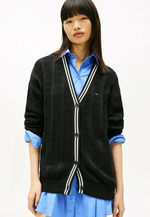 Black knit cardigan with textured pattern, featuring a V-neck, white striped accents, and black buttons. Worn over a blue button-up shirt.