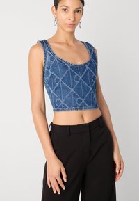 Top corto de mezclilla con escote redondeado, intrincado patrón de diamantes, tirantes anchos y dobladillo deshilachado. Combinado con pantalones negros de tiro alto.