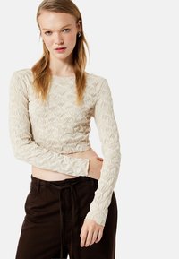 Cremefarvet langærmet top med tekstureret diamantmønster, cropped design og tætsiddende ærmer, parret med brune bukser med snor i taljen.