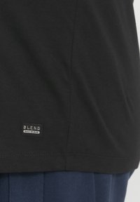 Crna majica od pamuka, s malom pravokutnom etiketom s tekstom "BLEND" i "BUILT IN BLUE" blizu donjeg ruba.