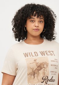Krēmkrāsas t-krekls ar uzrakstu "WILD WEST" brūnā krāsā. Iezīmēta rodeo grafika un Rietumu tēmas teksts. Īsas piedurknes un apaļa kakla izgriezums.