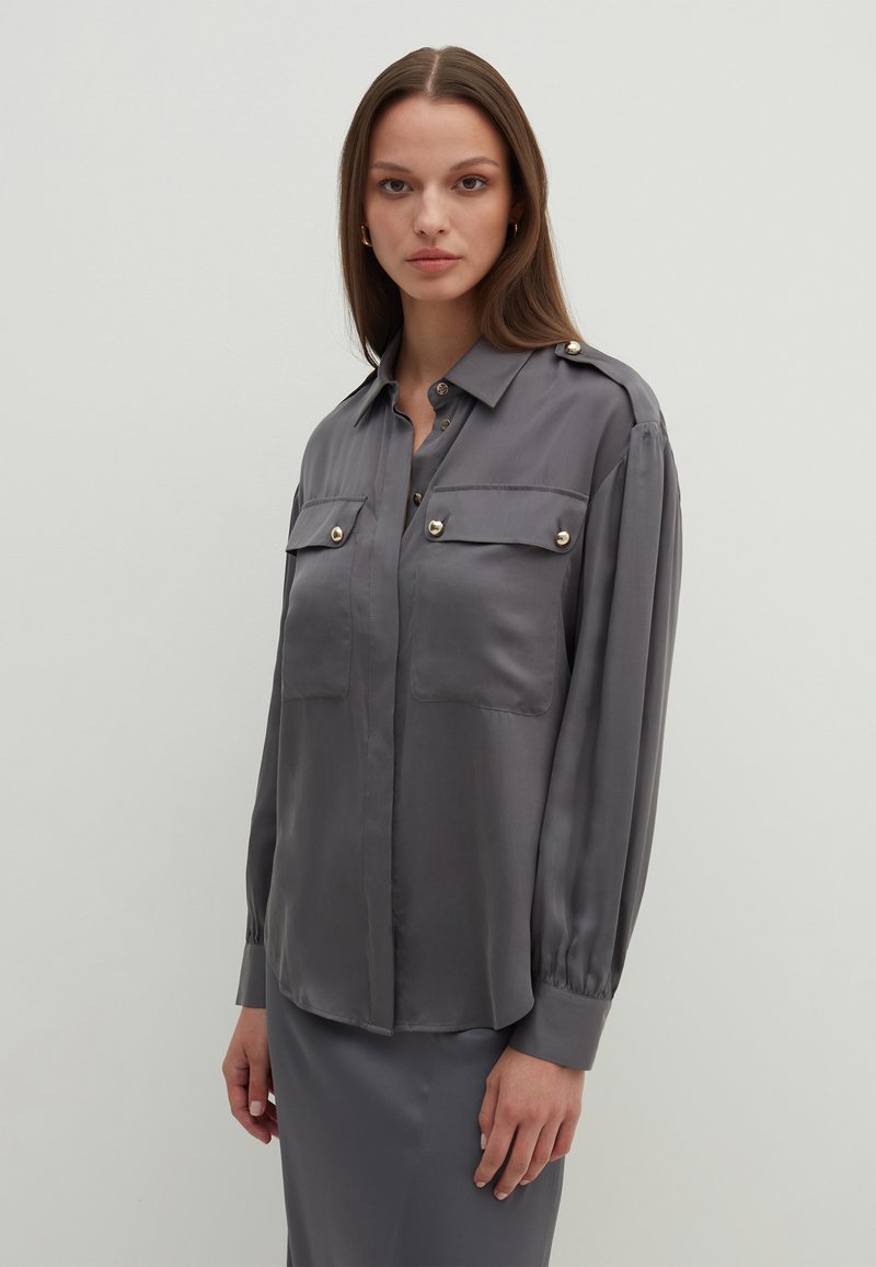 Camicia satin grigia con bottone frontale, due tasche sul petto, spalline sulle spalle e maniche lunghe con polsini abbottonati. Tessuto morbido e finitura liscia.