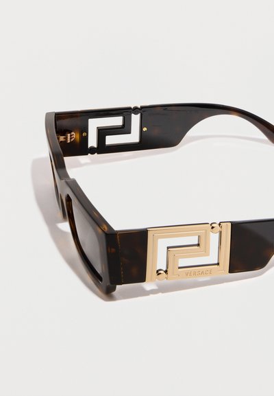 Καρέ ηλιοκαλύπτρες με σχέδιο από χελώνα, με φαρδιές πλευρές που διαθέτουν χρυσά γεωμετρικά ανοίγματα και το λογότυπο Versace σε λευκό φόντο.