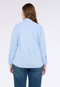 Světle modrá košile na knoflíky s dlouhými rukávy, klasickým límcem a mírně zakřiveným lemem. Úzké manžety mají zlaté knoflíkové detaily.