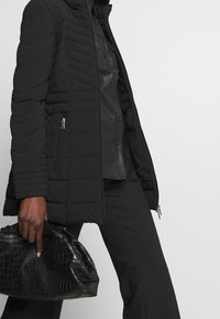 Giacca nera imbottita con colletto alto, tasche con zip e dettagli di cucitura a chevron, abbinata a una camicia nera in ecopelle e una borsa.