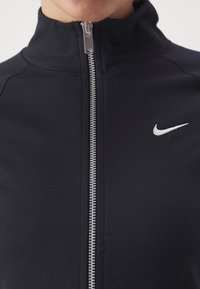 Svart zip-jacka med hög krage, som har en silverdragkedja och en liten vit Nike-logotyp på bröstet. Slät, elastisk tyg.