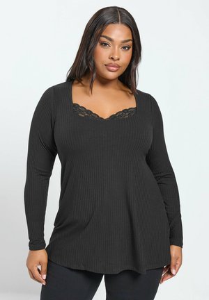 Mujer con el cabello liso y oscuro lleva una blusa negra de canalé de manga larga con detalles de encaje en el escote redondeado, posando contra un fondo liso.