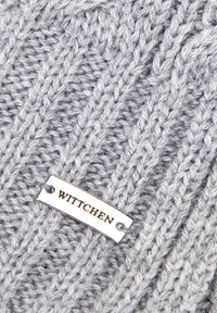 Grijze gebreide stof met een kabelpatroon en een zilveren label met de tekst "WITTCHEN" eraan bevestigd. De textuur is zacht en dik.