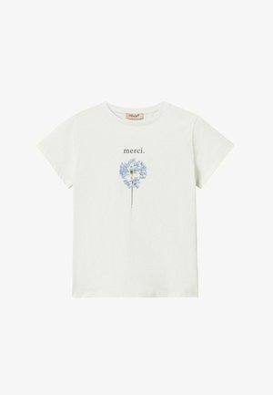 Joven mujer con camiseta blanca con gráfico, pantalones cortos azul marino texturizados, zapatos slingback blancos, sosteniendo un bolso azul marino con flecos y cadena dorada.