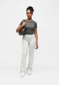 Femme aux cheveux bouclés portant un haut court gris foncé et un pantalon gris clair, avec un sac à main marron, debout sur un fond blanc.