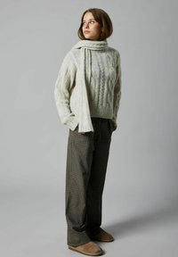 Chunky strik sweater i creme med høj hals, parret med brune ternede bukser og brune slip-on sko. Et tørklæde hænger over sweateren.