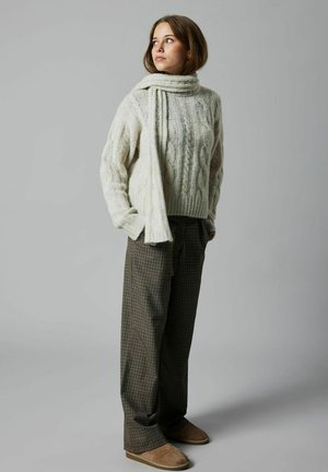 Jersey de punto grueso en color crema con cuello alto, combinado con pantalones marrón a cuadros y zapatos de slip-on marrones. Una bufanda cae sobre el jersey.