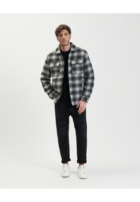 Giacca a camicia a scacchi bianco e nero con due tasche sul petto, indossata sopra un maglione nero, abbinata a pantaloni neri strappati e sneakers bianche.