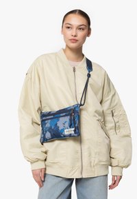 Sac bandoulière bleu tie-dye avec sangle ajustable, comportant des compartiments zippés et un patch logo. Porté sur une veste bomber beige.