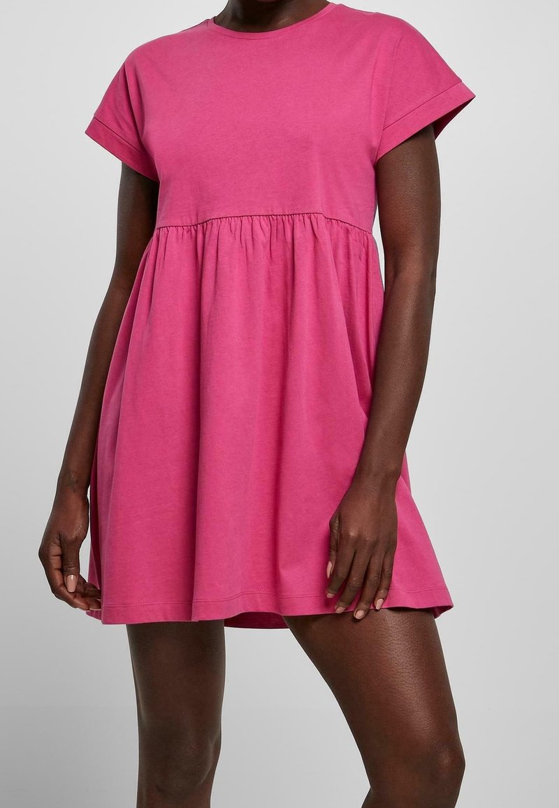 Femme portant une robe rose ample à manches courtes avec une taille froncée et un col rond, sur un fond uni clair.