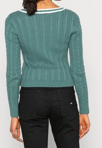 Ribbade tealtröja med långa ärmar och figursydd midja; har vita randiga detaljer vid halsringningen och flätmönster på ärmarna.