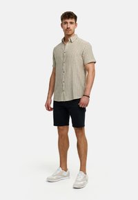 Short-sleeve button-up shirt in light beige with vertical stripes, paired with dark shorts and light sneakers. Smooth fabric, casual fit.