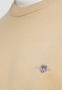 Sweat-shirt beige à col rond, avec un petit logo brodé en bleu, rouge et blanc. Tissu en coton lisse et doux.