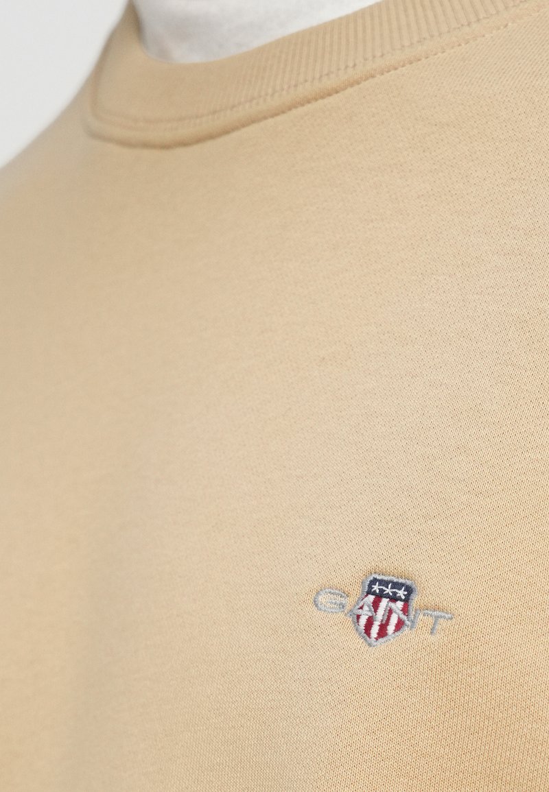 Sweat-shirt beige à col rond, avec un petit logo brodé en bleu, rouge et blanc. Tissu en coton lisse et doux.