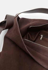 Sac fourre-tout en suede marron avec une bandoulière en cuir, comportant une poche zippée et un intérieur au toucher doux. Design à ouverture supérieure.