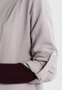 Długi rękaw w miękkim szarym materiale, z podwiniętym rękawem z bordowym akcentem dzianinowym i detalem z metalowego guzika w srebrnym kolorze.