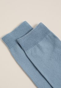 Modre bombažne nogavice z teksturirano površino in rebrastimi manšetami, rahlo prekrivajoče se, na nevtralnem ozadju.