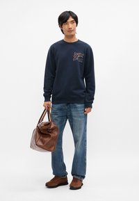 Sweatshirt navy avec un graphisme coloré, associé à un jean en denim bleu clair. Sac de voyage en cuir marron, et chaussures en daim marron avec des semelles noires.