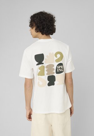 Personne aux cheveux bouclés portant un t-shirt blanc avec des formes abstraites beige, noire et verte et le mot « SPRING » imprimé dans le dos.
