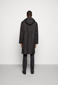 Impermeable negro, ligero, con capucha, mangas largas y corte recto. Cuenta con una abertura en la parte trasera para ventilación y una textura suave.