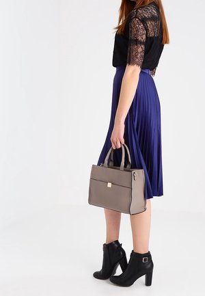 Femme portant un haut en dentelle noire, une jupe plissée violette, des bottines à talons noires, tenant un sac à main gris par ses poignées.